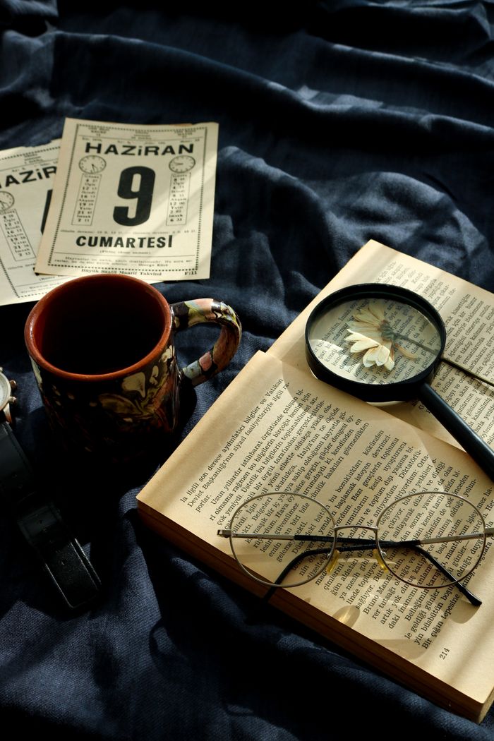 A nostalgic still life featuring a mug, open book, glasses, and magnifying glass on a dark cloth.